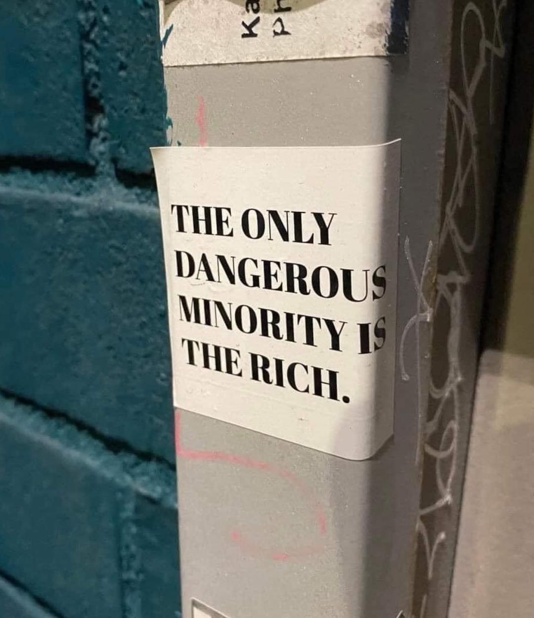 A street sticker pasted on a grey pole with graffiti around it reads in bold black capital letters: "THE ONLY DANGEROUS MINORITY IS THE RICH." The sticker contrasts against a teal brick wall in the background, making the political message stand out.