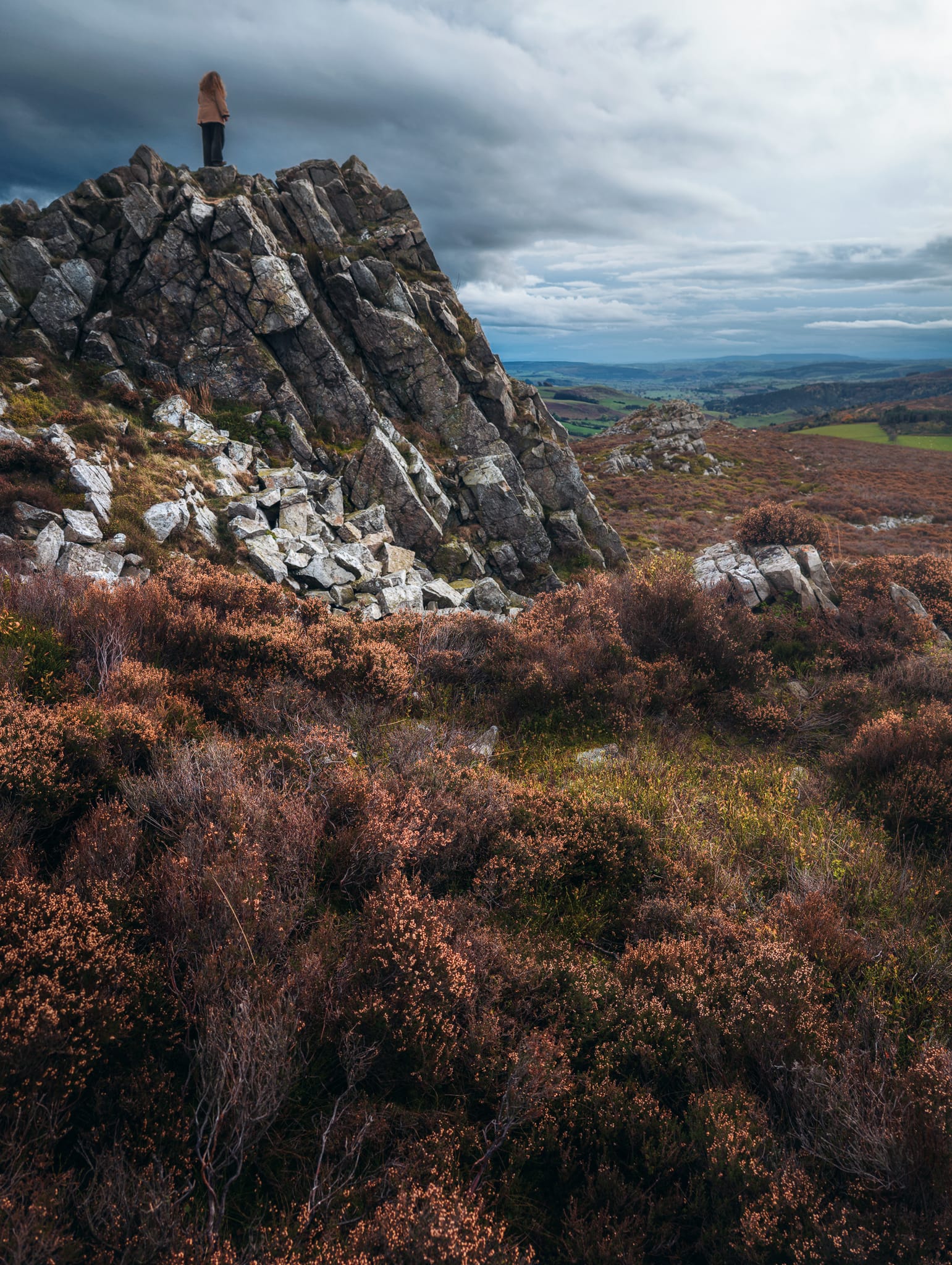 A lone person stands on top of a jagged outcrop of grey rock at Cranberry Rock on the Stiperstones, looking out over a broad rural landscape under a heavy sky. The crags rise sharply from heather moorland that fills the foreground with russet and earthy tones. Beyond the main ridge, rolling hills and patchwork fields stretch into the distance with muted autumn colours. Low cloud and soft, diffused light give the scene a moody and atmospheric feel.